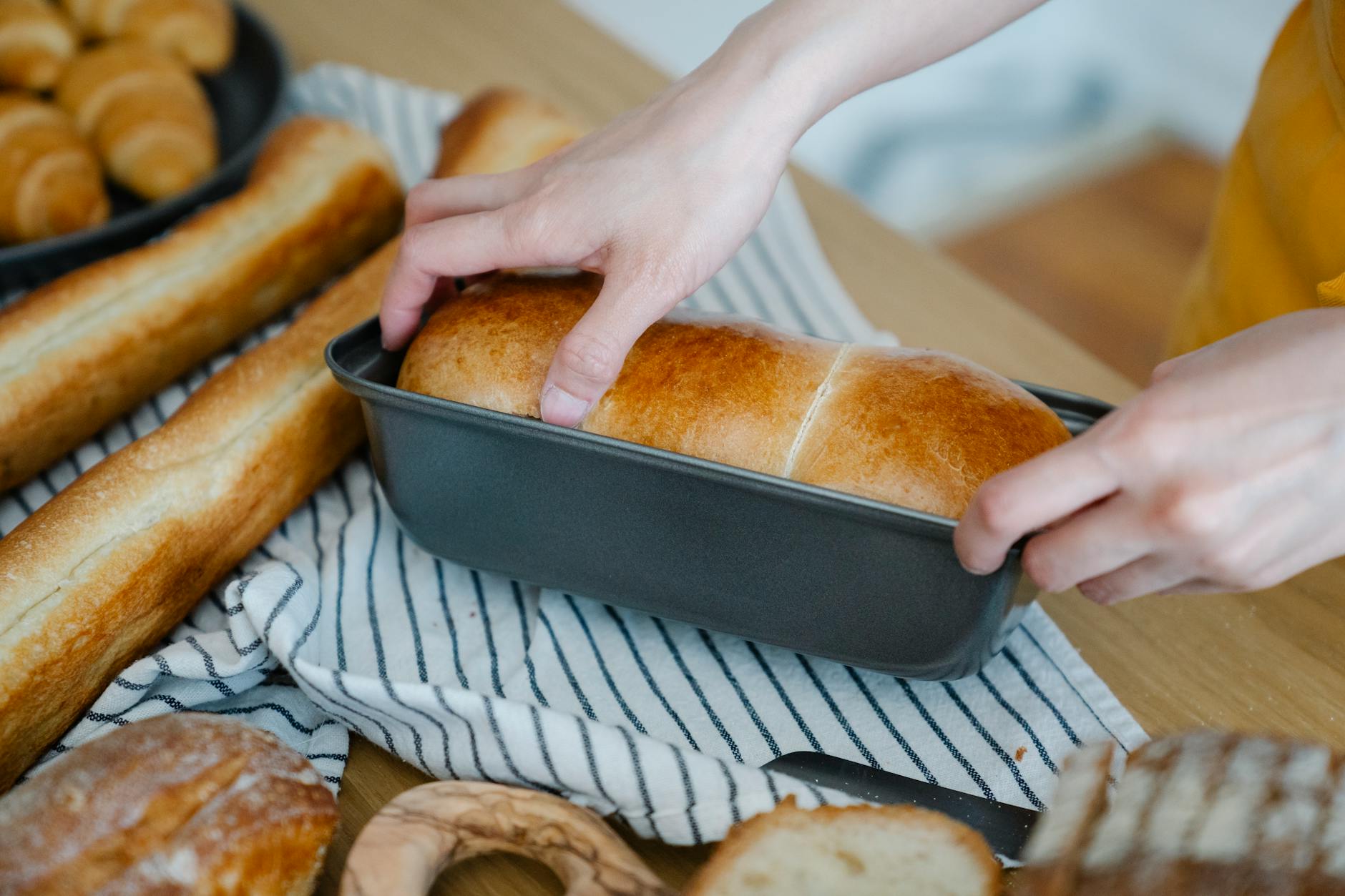 Dłonie trzymają świeżo upieczony chleb pszenny w foremce, obok inne bochenki