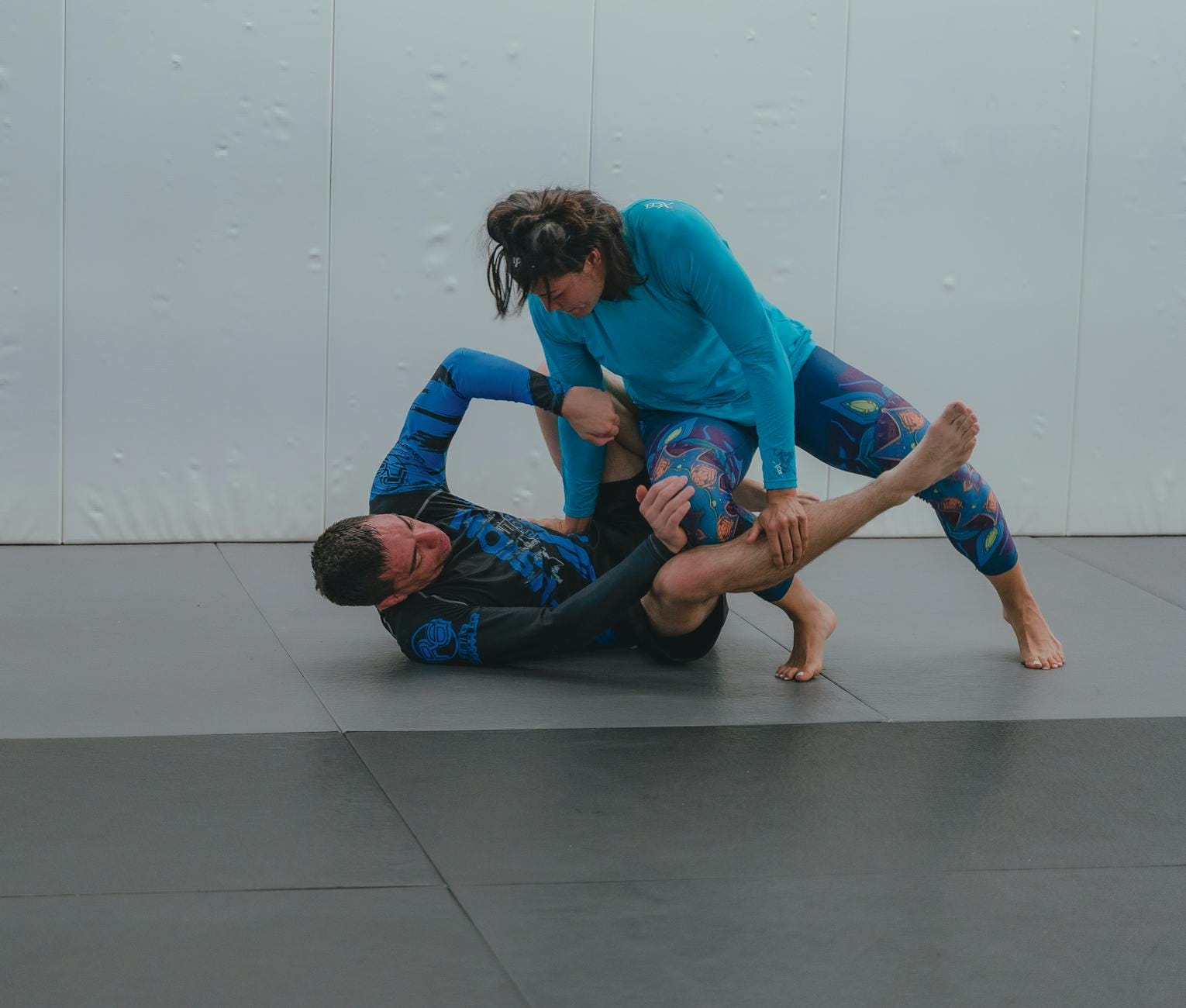 Dwóch dorosłych trenuje brazylijskie jiu-jitsu na macie w sali